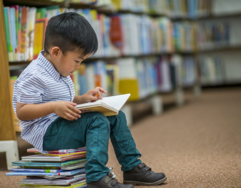 A young Asian boy is indoors in his elementary school library. He is reading a storybook while sitting on a stack of books.